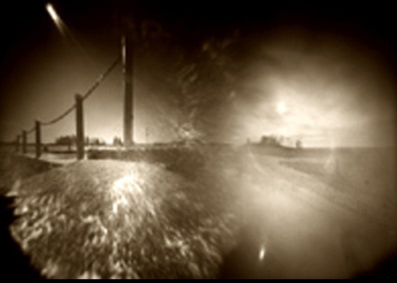 Sepiafarbenes Lochkamerabild aus zwei Belichtungen: links ein Weidezaun und ein Telegrafenmast vor tiefstehender Sonne, rechts eine gleißend helle Ebene, beide Hälften gehen ineinander über.